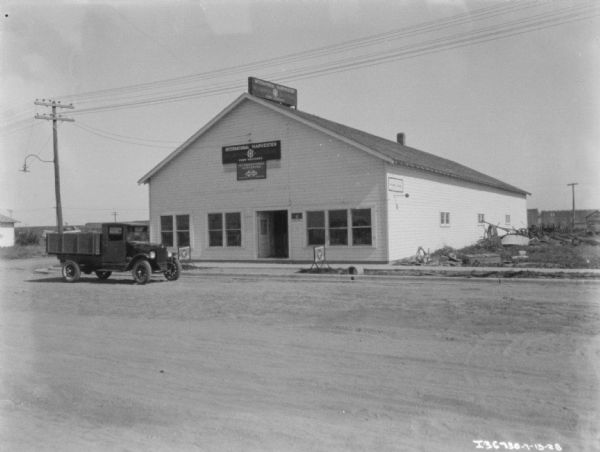View across road towards a truck near a building with a sign that reads: "International Harvester McCormick-Deering Farm Machines."