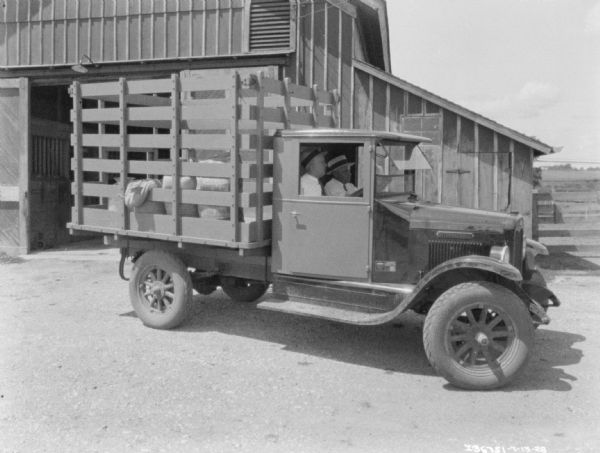 View towards two man sitting in the cab of a truck. The truck bed has a high stake body for livestock, and is parked in front of a barn.