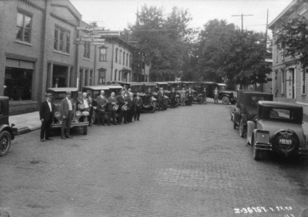 View down cobblestone street towards a group of men posing with a line of trucks, which are parked in front of a brick building.
