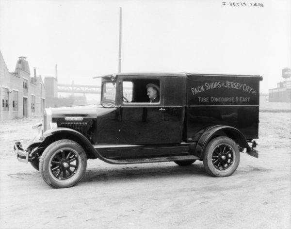 View towards a man sitting in the driver's seat of a delivery truck. The sign on the side of the truck reads: "Pack Shops of Jersey City Inc. Tube Concourse 9 East." Industrial buildings are in the background.