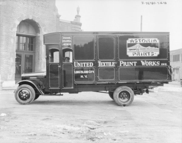 View towards the driver's side of a large delivery truck, parked near a brick building with a sign for "Motor Trucks" above the arched entrance. Two people are sitting inside the cab of the truck. The sign painted on the side of the truck reads: "United Textile Print Works Inc." and "Astoria Prints."