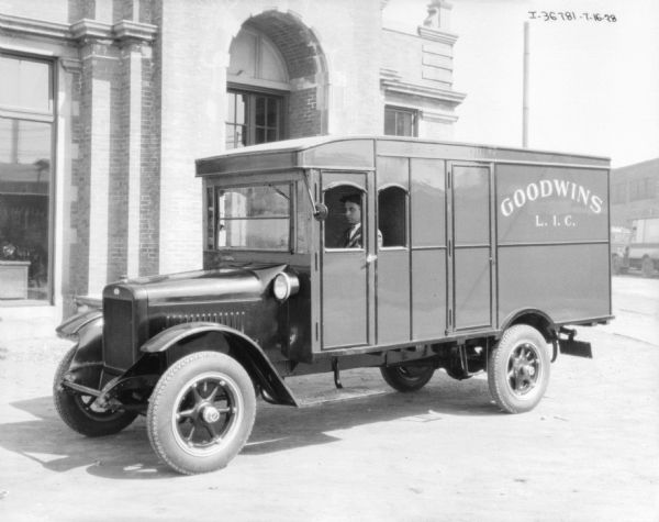 View towards the driver's side of a delivery truck. A man is sitting in the driver's seat, and the sign painted on the side reads: "Goodwins L.I.C." Behind the truck is a brick building with an arched entrance.