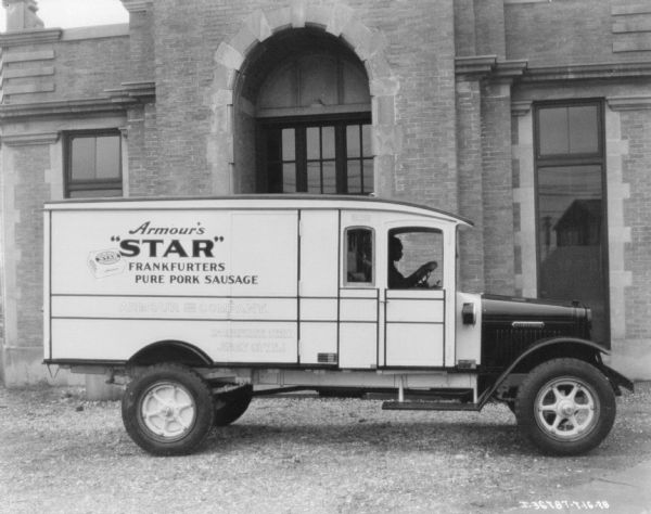 View towards the passenger side of a truck parked in front of a brick building with an arched entrance. A man is sitting in the driver's seat, and the sign painted on the side of the truck reads: "Armour's 'Star' Frankfurters Pure Pork Sausage."