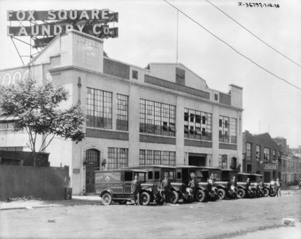 View across road towards a group of men standing with a fleet of delivery trucks in front of a large building. A large electric sign on top of the building reads: "Fox Square Laundry Co."
