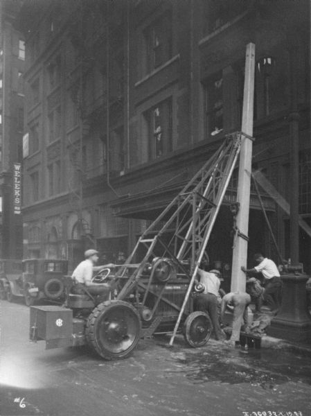 View down street towards a man driving a McCormick-Deering tractor to install a telephone pole on a city block. Other men are standing at the front of the tractor to help position the pole.
