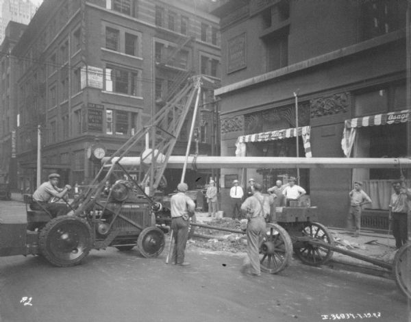 View down street towards a man using a McCormick-Deering tractor with a hoist to install a lamppost on a sidewalk.
