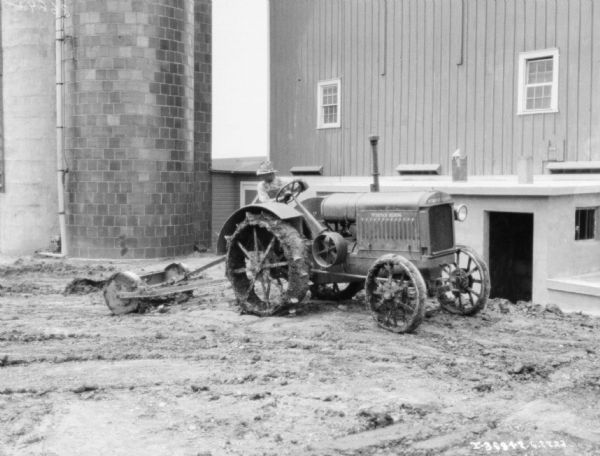 A man is driving a McCormick-Deering tractor pulling an agricultural implement in the mud in a barnyard near a barn and a silo.