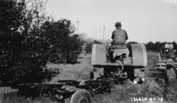 Right side view of a man using a tractor to pull a disk harrow in a field or orchard. An automobile is parked in the background on the right.