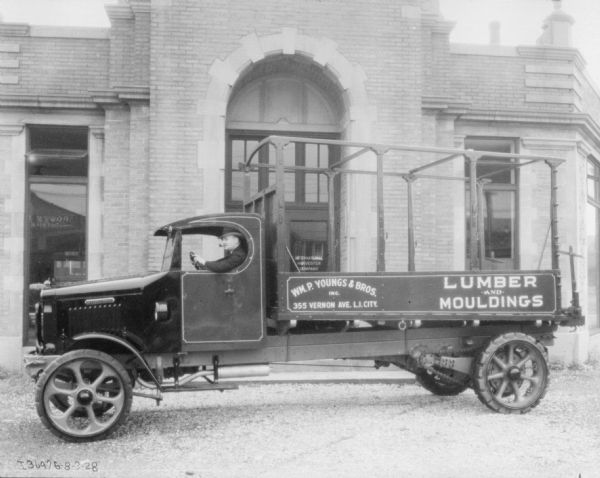 A man is sitting in the driver's seat of a truck smoking a pipe. The truck is parked in front of an arched entrance, which has a sign for International Harvester Company on the front door. The sign painted on the side of the truck reads: "Wm. P. Youngs & Bros., 355 Vernon Ave., L.I. City. Lumber and Mouldings."