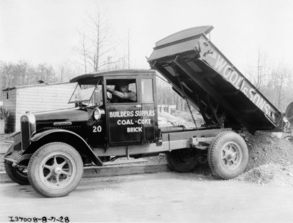 A man is sitting in the driver's seat of a dump truck. The back of the truck is tipped back into a pile of material. The sign painted on the side of the truck reads: "Builders Suppplies, Coal • Coke, Brick, G?.D. Wood & Sons."