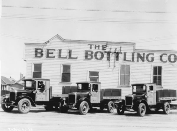 Three men are sitting in the driver's seats of three delivery trucks. They are parked in front of a large building with a sign painted on it that reads: "The Bell Bottling Co." The signs painted on each of the driver's doors reads: "Grape Squeeze."