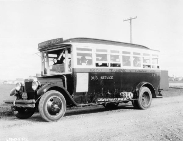 View towards the left side of a bus. People are sitting inside the bus which is parked on an unpaved road. The sign painted on the side reads: "Royal Transfer & Storage Ltd." A sign above the front window reads: "Winnipeg - Teulon." There is a Manitoba license plate on the front of the bus.