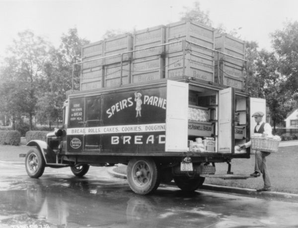 A man is standing at the back of a bakery delivery truck. He is holding a basket, and the back doors of the truck are open, showing shelves of bread and other bakery items. Large crates of bread are also stacked on top of the truck enclosed by a metal railing and chains. The sign painted on the side of the truck reads: "Speirs Parnell Bread, Serve Two Kinds of Bread Every Meal, Bread, Rolls, Cakes, Cookies, Doughnuts." There is a Manitoba license plate on the back of the truck.