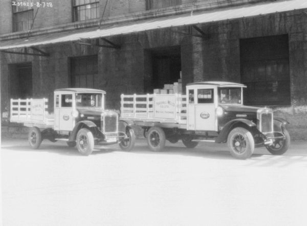 Two trucks with stake bodies are parked along the side of the loading dock of a large, brick building. There is a roof above the trucks. The signs on the side of the trucks reads: "Marshall-Wells Co. Ltd. Wholesale Hardware."