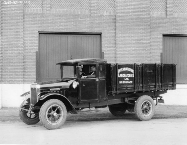 View towards a man sitting in the driver's seat of a truck parked in front of a large, brick building. The sign painted on the side of the truck bed reads: "International Laboratories Ltd. St. Boniface."