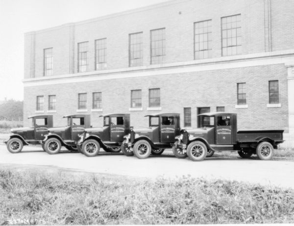 View towards five trucks parked in a row at an angle. Two men are sitting in the driver's seats of two of the trucks. In the background is a large, brick building. The signs painted on the driver's side doors read: "City of Winnipeg, Waterworks."