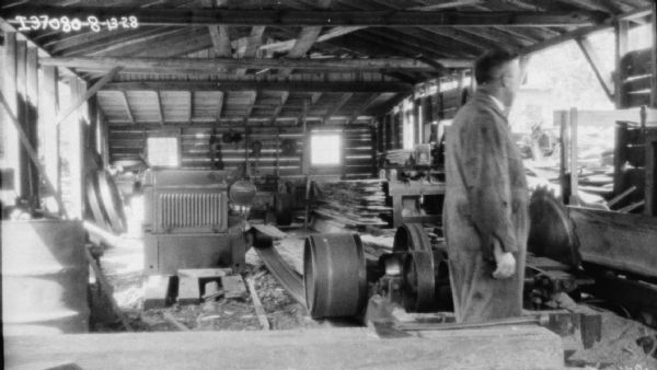 Interior view of a sawmill with an engine powering the saw. A man is standing in the right foreground near the sawblade.