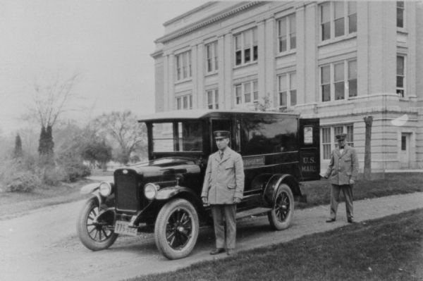 Two men in uniform are standing with a U.S. Mail truck. They are parked in the driveway with a large building behind them.