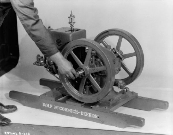 Assembly maintenance. View of a person (mostly out of frame) whose hand is holding a wrench making adjustments on a machine. Sign painted on wood frame under machine reads: "1 1/2 H.P. McCormick-Deering."