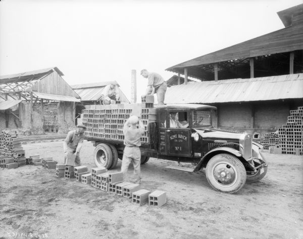 Four men are loading bricks onto the back of a truck. A man is sitting in the passenger seat of the cab. The sign on the passenger door reads: "Acme Brick Co. Ltd. No. 1."