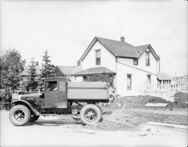 View towards a truck parked near a house, with a man sitting in the driver's seat. The side yard is a construction site. A man is working in the yard among piles of dirt. There is a horse pulling a wagon on the far left.
