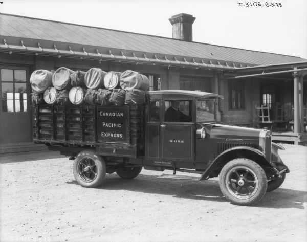 View of the passenger side of a truck loaded with bundles. The truck has a stake body, and a man is sitting in the driver's seat. A sign on the side of the truck reads: "Canadian Pacific Express." Just behind the truck is a long, one-story building with carts on a loading dock on the right. Under the passenger door is a sign that reads: "Made By International Harvester Company of Canada, Ltd., Hamilton, Ontario."