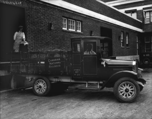 View of the passenger side of a truck with a stake body, with a man is sitting in the driver's seat. A sign on the side of the truck bed reads: "Canadian National Railways." The truck is backed up to a loading dock in a brick building on the left, and a man wearing an apron is standing on the loading dock.