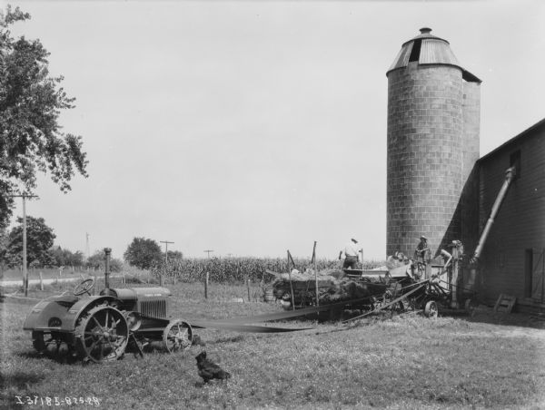 View across barnyard towards men using a thresher near a barn. A tractor in the yard on the left is belt-driving the thresher to fill the haymow. There is a silo near the barn.