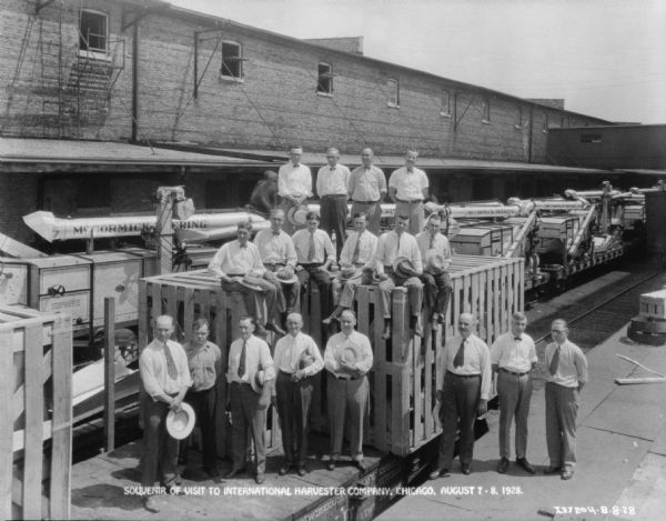Elevated view of a group of men posing on and around a machine in a crate. Behind the group are agricultural machines, and a long, brick building. Caption on bottom reads: "Souvenier of visit to International Harvester Company, Chicago, Aug. 7-8, 1928."