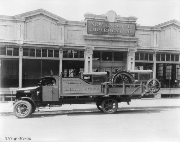 View across street towards a two men in the cab of a truck transporting two McCormick-Deering tractors parked in front of the New England Implement Co. building. A man is sitting in the driver's seat of the delivery truck, which has a sign on the side of the truck bed that reads: "McCormick-Deering Industrial Tactors Operating Equipment, New England Implement Co."