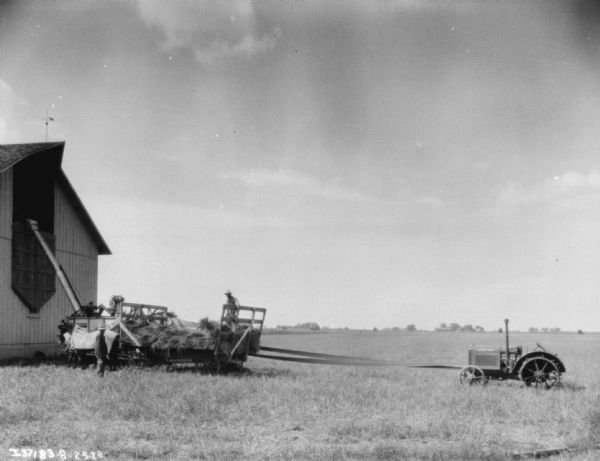View towards men using a thresher to fill a haymow in a barn. A tractor is belt-driving the thresher.