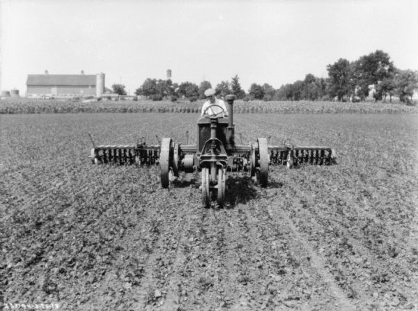 Front view of a man driving a Farmall tractor pulling rotary hoes in a field. Farm buildings are in the background.