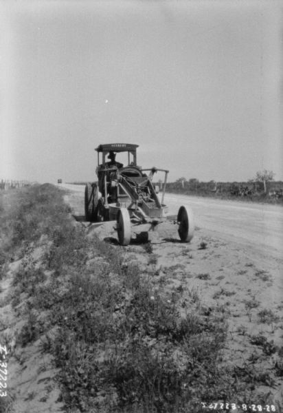 View down side of road towards a man using a grader. The sign on the front of the roof reads: "Gilbert."