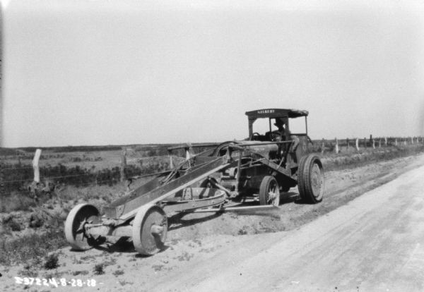 Three-quarter view from front left towards a man using a grader on the side of a road.