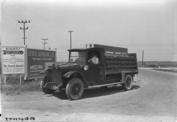 Two men are sitting in the cab of a beverage delivery truck, which is parked on a road. The sign on the truck over crates of bottles reads: "Drink Delicious Orange Crush." In the background are billboards. One reads, in part: "Right! To the Valley and Laredo."