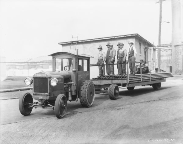 A group of men are standing and sitting on a trailer being pulled by a man driving a McCormick-Deering tractor. Buildings are in the background.