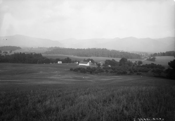View down hill towards farm buildings in a valley. Hill and mountains are in the distance.