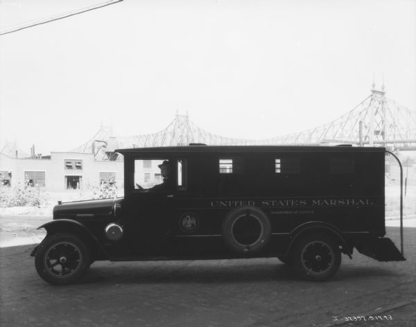 Left side profile view of a man sitting in the driver's seat of a truck. The sign painted on the side reads: "United States Marshal, Department of Justice." In the background are buildings, and a large bridge.
