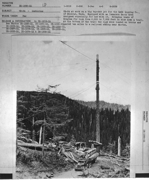 View towards man using a TD-24 to load logs onto a truck. Mountains are in the background. Subject: "TD-24 — Lumbering." Where Taken: "Pac." Information with photograph reads: "TD-24 at work on a big try-out job for the Ladd Logging Co., of Mineral, Wash. Equipped with an Isaacson Karry arch designed especially for use with it. Bringing loads of Douglas fir logs from 6,000 to 7,000 feet in size down a trail. At the bottom of the trail the logs were loaded on trucks and hauled ten miles to a railroad siding near Morton."