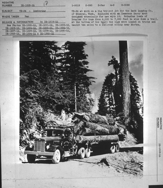 View down road towards a truck loaded with logs. The sign painted on the driver's side door reads: "Lloyd E. Larson, Arcata, Calif." Subject: "TD-24 — Lumbering." Where Taken: "Pac." Information with photograph reads: "TD-24 at work on a big try-out job for the Ladd Logging Co., of Mineral, Wash. Equipped with an Isaacson Karry arch designed especially for use with it. Bringing loads of Douglas fir logs from 6,000 to 7,000 feet in size down a trail. At the bottom of the trail the logs were loaded on trucks and hauled ten miles to a railroad siding near Morton."