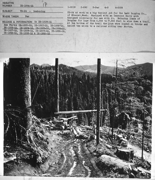 View down trail towards a man using a TD-24. Mountains are in the background. Subject: "TD-24 — Lumbering." Where Taken: "Pac." Information with photograph reads: "TD-24 at work on a big try-out job for the Ladd Logging Co., of Mineral, Wash. Equipped with an Isaacson Karry arch designed especially for use with it. Bringing loads of Douglas fir logs from 6,000 to 7,000 feet in size down a trail. At the bottom of the trail the logs were loaded on trucks and hauled ten miles to a railroad siding near Morton."