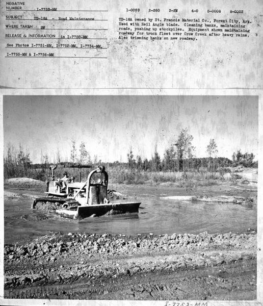 View towards a man driving an TD-18A in water. Subject: "TD-18A — Road Maintenance." Where Taken: "SE." Information with photograph reads: "TD-18A owned by St. Francis Material Co., Forest City, Ark. Ussed with Heil Angle blade. Cleaning banks, maintaining roads, pushing up stockpiles. Equipment shown maintaining roadway for truck fleet over Crow Creek after heavy rains. Also trimming banks on new roadway."