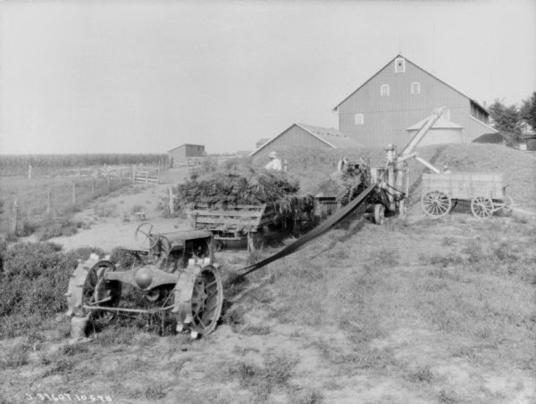 Slightly elevated view of men working in a farmyard with a tractor belt-driving threshing machinery. One man is standing on a wagon loaded with hay, and another person is in a wagon near the thresher, which is piling up the threshed hay into large piles near farm buildings.