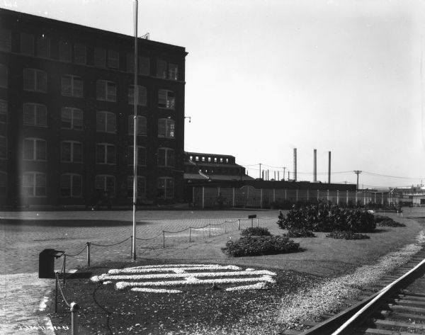 View of the International Harvester plant. In the foreground is a fenced area, with the IHC logo on ground near a flagpole and flower beds. Factory buildings are in the background. Railroad tracks are on the right.