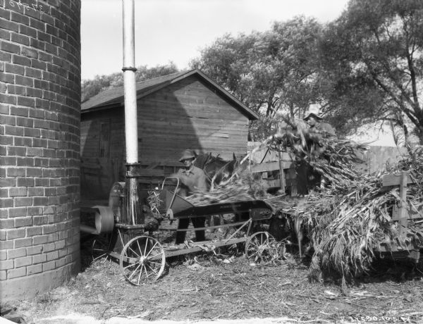 Close-up of a man using an ensilage cutter to fill a silo. Another man is standing in a horse-drawn wagon the right.
