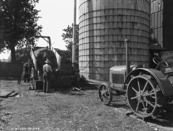 A man is using a tractor to belt-drive an ensilage cutter to fill a silo. The man is working at the back of a horse-drawn wagon.