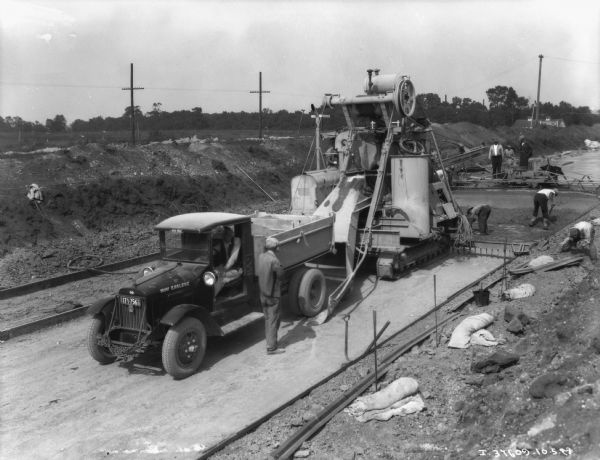 Elevated view of a group of men working on highway construction. A man is sitting in the driver's seat of a truck, with a machine with a chute directly behind it. A group of men are working leveling the asphalt on the road behind the truck.