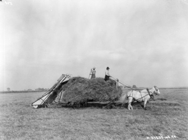 Right side view of two men on a horse-drawn wagon with a hay loader on the back.