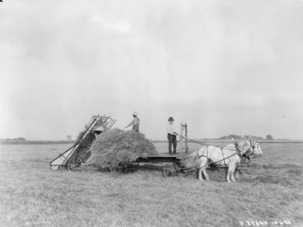 View across field towards two men on a horse-drawn wagon with a hay loader on the back.