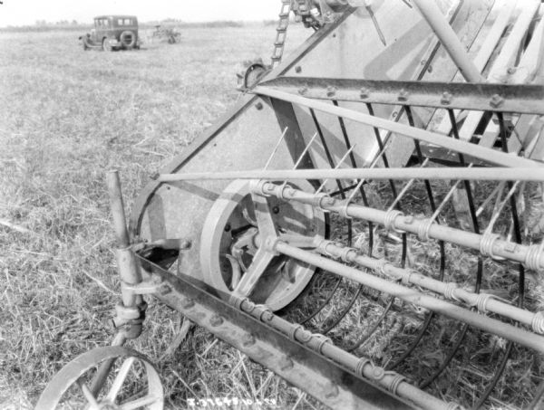 Close-up view of a section of a hay loader parked in a field. There is an automobile parked in the background.
